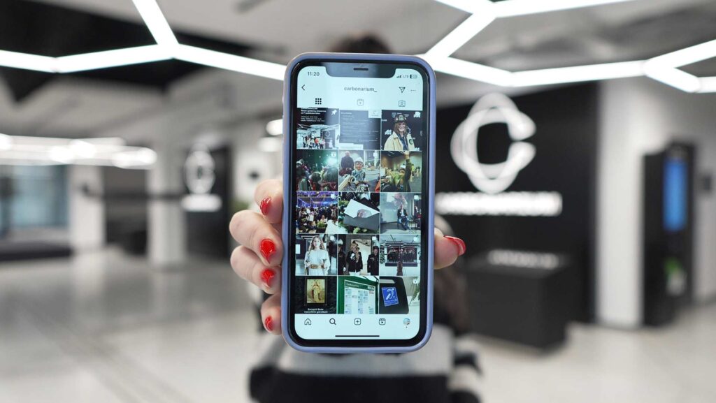 Kobieca dłoń wyciągnięta w stronę obiektywu. W dłoni telefon z włączonym profilem Carbonarium na platformie Instagram.